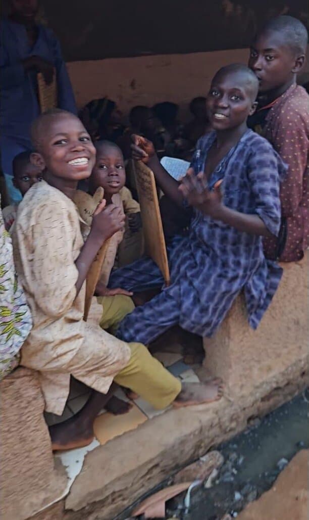 Two smiling boys holding a wooden Quranic study tablet at an outdoor school in Kaduna